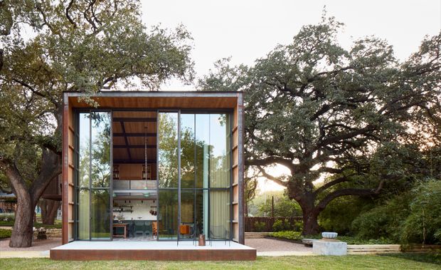 A compact modern glass pavilion set within a landscaped garden, framed by mature spreading trees. The structure has a rectangular timber-clad facade, extensive floor-to-ceiling glazing with slender dark frames, and a flat roof. A small interior workspace or living area is visible through the glass, with a simple concrete platform at the front.
