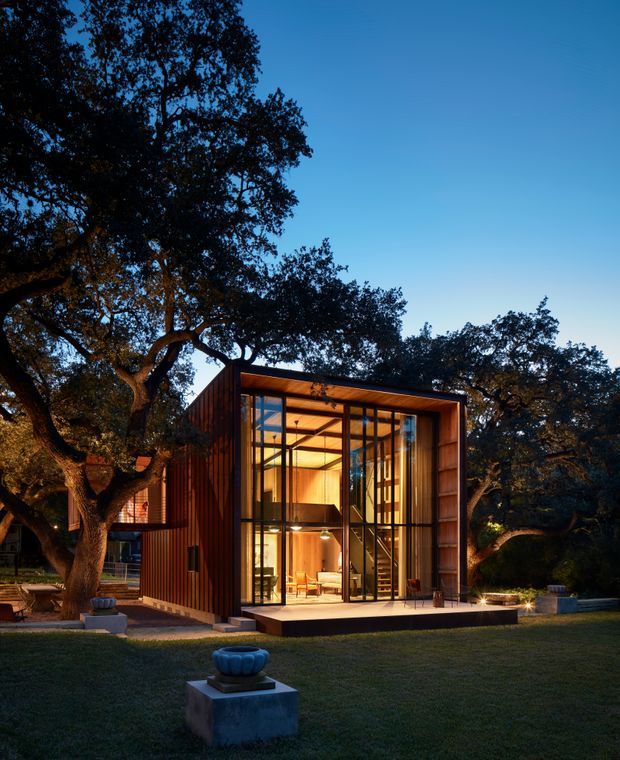 A compact modern pavilion or small house set within a mature wooded garden at dusk. The building features a strongly rectilinear form, dark vertical timber cladding, extensive floor-to-ceiling glazing, a projecting flat roof, and a warm timber-lined interior visible through the glass. A small raised terrace and sculptural planters sit in front, while large established trees frame the structure.