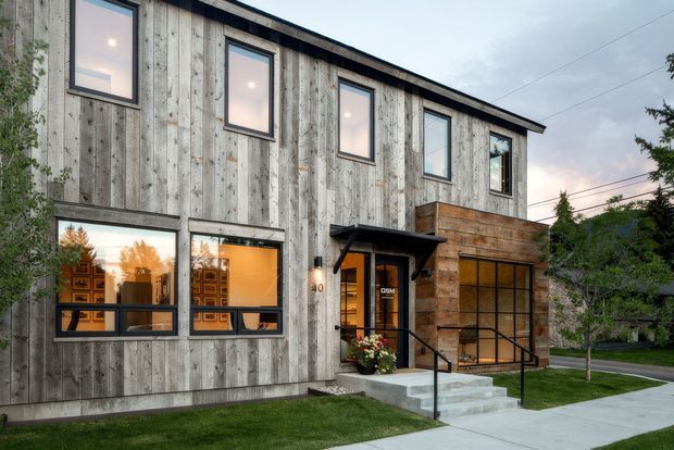 Two-storey contemporary building with a long, simple rectangular form. The facade combines weathered grey vertical timber cladding with a contrasting warm timber-clad entry volume, dark metal-framed windows, and a small projecting entry canopy. Large ground-floor glazing reveals warm interior lighting and patterned screens or joinery. A concrete entry stair, paved footpath, lawn, and mature trees frame the exterior.