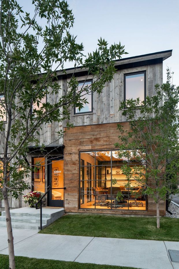 A compact two-storey contemporary building viewed from the landscaped front walkway. The facade combines weathered vertical timber cladding with horizontal natural timber boards, dark window and door frames, and a simple low-pitched roof. Floor-to-ceiling glazing at ground level reveals a warmly lit interior workspace or living area, while young trees, lawn, concrete paving, and a small entry stair frame the exterior.
