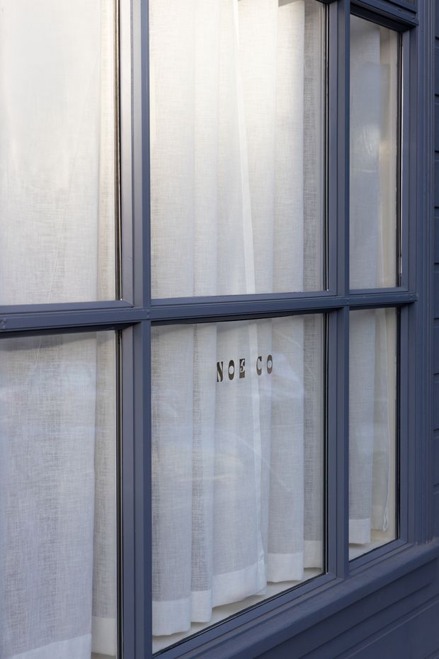 Close-up exterior view of a dark blue painted window and door-style glazing assembly with multiple rectangular panes, set within matching blue cladding. Sheer white curtains are visible behind the glass, with subtle lettering on the interior fabric. The image focuses on the facade envelope and window framing rather than an identifiable interior room.