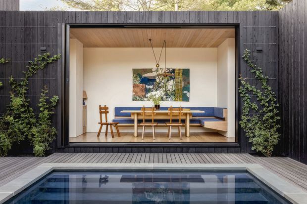 A contemporary poolside outdoor view looks into a compact dining and entertaining room through a large glazed opening. The room has a built-in blue upholstered banquette, a light timber dining table and chairs, a colorful abstract artwork, and a warm timber-lined ceiling. The surrounding exterior is clad in dark vertical timber boards with climbing greenery, and a paved timber deck borders the swimming pool in the foreground.
