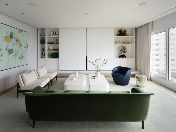 Bright, refined contemporary living room with a large green upholstered sofa in the foreground, two cream lounge chairs, a white rectangular coffee table, and a sculptural navy armchair. The room has pale wood flooring, white walls and ceiling with recessed spotlights, full-height built-in shelving and cabinetry, sheer and heavier curtains, and expansive corner glazing overlooking neighbouring apartment buildings. Minimal artwork and curated decorative objects add colour and texture.