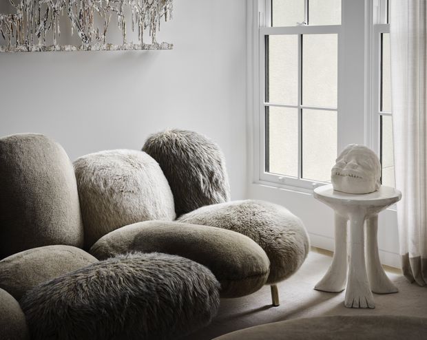 A softly lit, monochromatic sitting area with a sculptural, heavily textured upholstered lounge chair or sofa in grey and taupe tones. A tall multi-pane window with white trim fills the right side, while a small sculptural pedestal table supports a pale decorative object. A textured abstract artwork is partially visible above the seating, and sheer curtains frame the window.