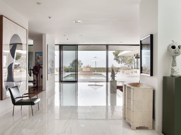 Bright, spacious contemporary interior with a polished white marble floor, white walls and ceiling, and full-height dark-framed glazing opening toward an outdoor terrace and pool area. The room includes minimalist cabinetry, sculptural artwork, a modern lounge chair, and a large framed wall-mounted display. The open-plan space has a refined, gallery-like atmosphere and strong indoor-outdoor connection.