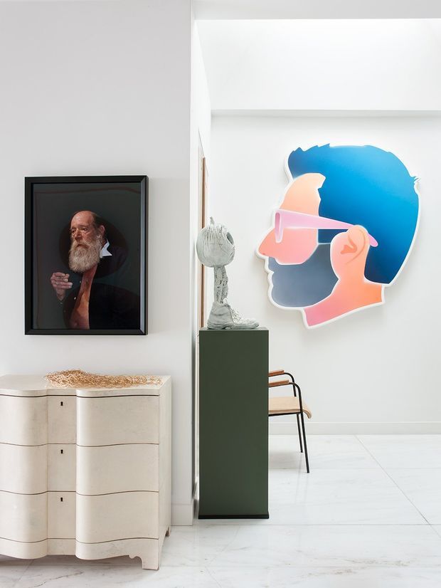 Bright, curated interior vignette with white walls and pale flooring. A sculptural cream-coloured chest of drawers sits beneath a framed portrait, while a dark green pedestal supports a small sculptural object. A large colourful graphic wall artwork dominates the adjoining white wall, with part of a dining chair visible behind the pedestal. The space has a clean, gallery-like presentation with eclectic contemporary art and furnishings.