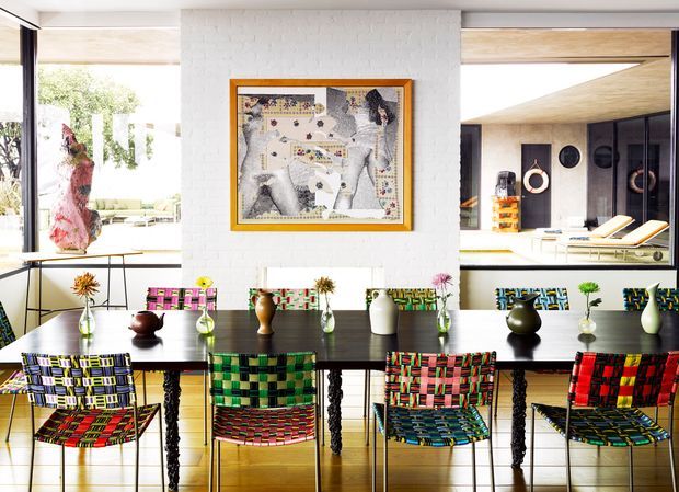 Bright contemporary dining area with a long dark dining table and colorful woven chairs, set against a white-painted brick feature wall. Large internal windows open the space to adjacent living and lounge areas, while an artwork, sculptural pendant, and vases add eclectic colour and texture.