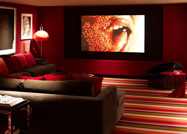 A dark, dramatic home cinema or media room with deep red built-in wall panelling, a large wall-mounted screen, multiple brown and red sofas, a black lounge chair, and a bold multicoloured striped carpet. Warm low-level lighting and saturated red finishes create a theatrical atmosphere.