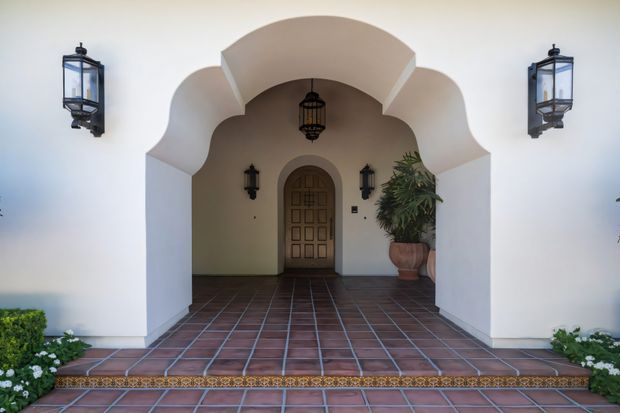 Front entry porch of a stucco residence with a deeply recessed, sculpted arched portico. The entrance features a paneled timber door, dark metal lanterns, a hanging pendant lantern, terracotta tile flooring, and potted greenery. The symmetrical white facade and decorative arch forms create a Mediterranean or Spanish-influenced character.
