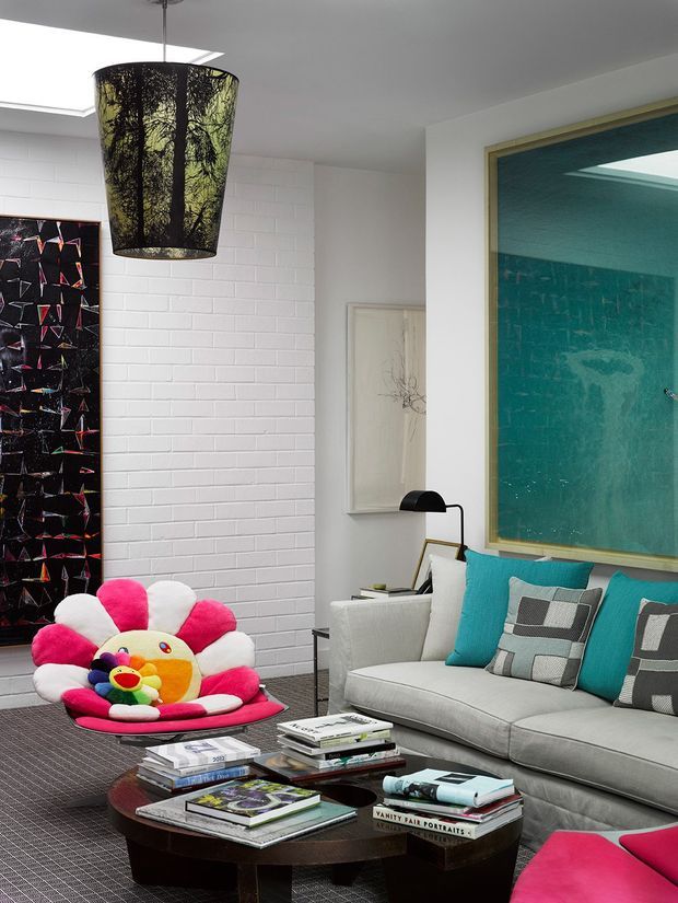 Eclectic contemporary living room with a light grey sectional sofa, vivid turquoise cushions, a dark round coffee table stacked with books, and a playful pink-and-white flower-shaped children's chair. The room features a white-painted brick wall, large framed artwork, a dramatic forest-pattern pendant light, a skylight, and a large turquoise-toned framed artwork or glazed opening.