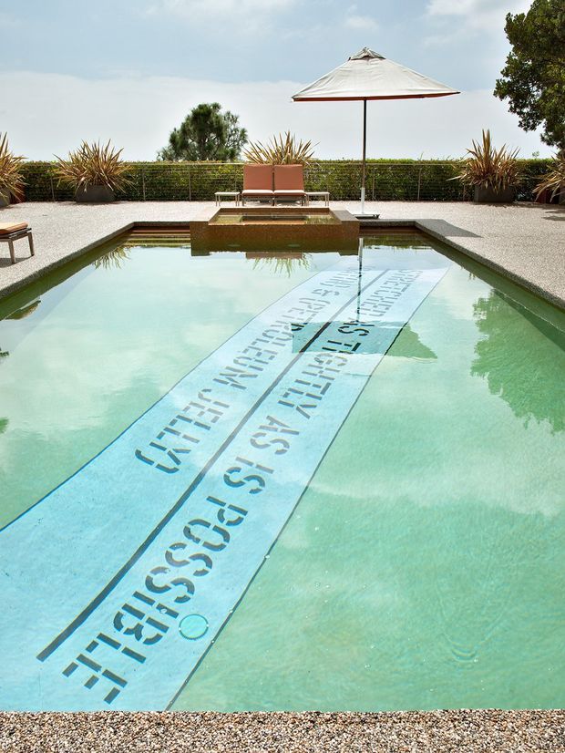 Outdoor swimming pool with a long typographic tiled lane or inset artwork running through the water. The pool is surrounded by pale gravel paving, clipped hedging, ornamental grasses, sun loungers and a large white parasol, creating a bright resort-like setting.