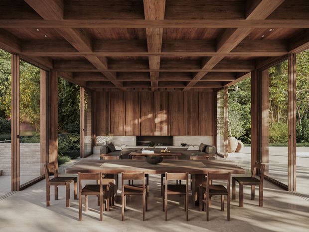 A warm, timber-lined open-plan dining and living space with a large solid wood dining table and chairs in the foreground. The room features extensive floor-to-ceiling glazing, exposed timber post-and-beam construction, a coffered timber ceiling, timber wall panelling, and a built-in kitchen or bar area at the rear. Lush garden planting is visible through the open glazing.