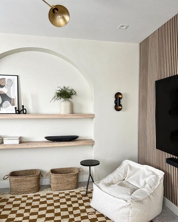 A refined contemporary living room or media area with a sculptural arched recessed display niche, pale wood floating shelves, woven baskets, a white textured lounge chair, checkered rug, and black wall-mounted television. A vertical timber slat feature wall adds warmth and texture, complemented by brass-toned lighting and wall sconces.