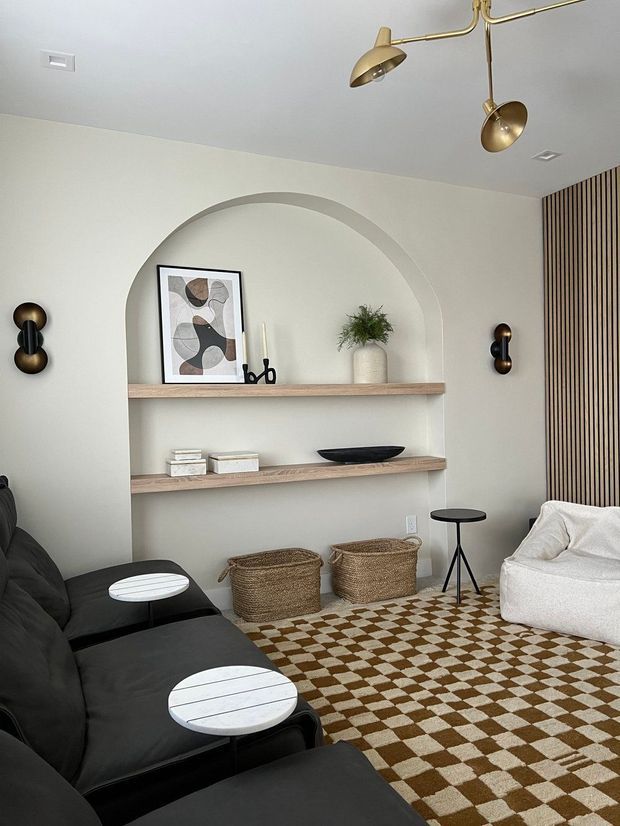 A refined contemporary living room with a built-in arched shelving niche, pale neutral walls, light timber shelves, sculptural decor, a black sectional sofa, cream lounge chair, brass wall and ceiling lights, and a brown-and-cream checkerboard rug. Vertical timber battens add texture along the right wall.