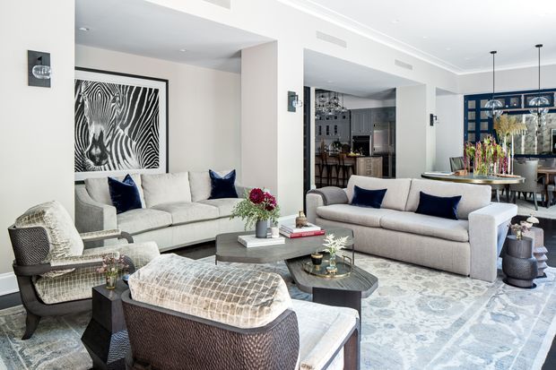 Bright, refined living room with two light upholstered sofas, sculptural armchairs, dark blue cushions, layered coffee tables, and a large pale patterned rug. A large black-and-white artwork anchors the seating area, while an open kitchen and dining space are visible beyond. White walls, built-in architectural openings, recessed ventilation, and dark-framed glazed cabinetry create a polished contemporary interior.