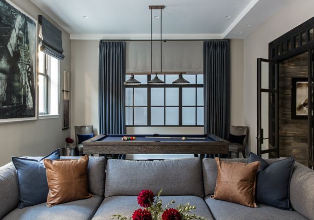 A refined living room with a large grey sectional sofa in the foreground, accented by navy and cognac leather cushions. A billiards table sits centrally beneath a black-framed industrial-style window, with dark curtains and three pendant lights above. The room features neutral painted walls, black-framed internal glazing, timber-look wall panelling, and restrained contemporary furnishings.