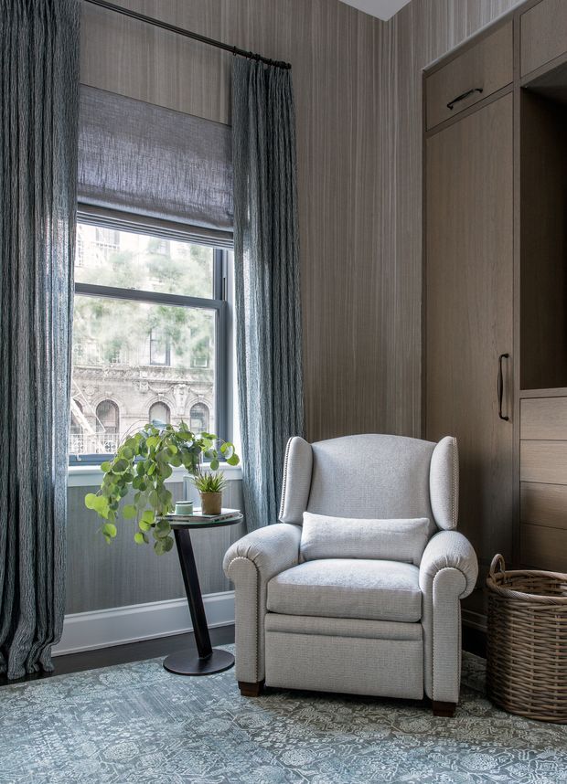 A refined reading nook with a light upholstered wingback armchair positioned beside a tall window. The room features vertically textured taupe wallcovering, dark blue-grey curtains, a patterned blue-grey area rug, a small black side table with potted plants, and built-in dark timber cabinetry with drawers and open shelving. A woven basket sits beside the chair. An ornate stone building is visible outside the window.