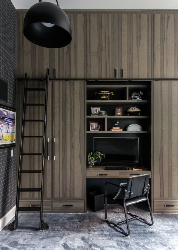 A dark, sophisticated built-in study and storage wall with tall vertically grained timber cabinetry, integrated drawers, open display shelving and a recessed television. A black metal library ladder stands beside the cabinetry, while a compact desk with computer monitor and a black leather cantilever chair forms a home office nook. The room includes a large black dome pendant light, dark small-format tiled wall, framed sports imagery and a deep grey patterned rug.