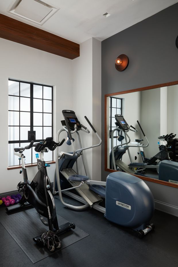 A compact home gym with multiple cardio machines, including treadmills, an elliptical trainer and an upright exercise bike. The room has dark rubber flooring, a charcoal feature wall, white walls and ceiling, a large black-framed multi-pane window, a full-width timber-framed mirror, a timber ceiling beam, and a wall-mounted copper-toned light. Dumbbells and other exercise equipment are arranged along the floor.