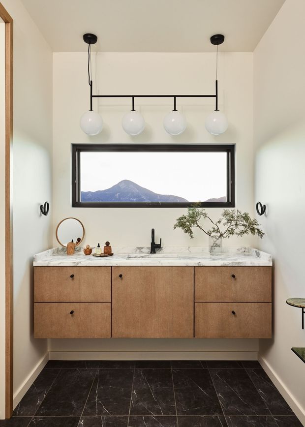 A refined bathroom vanity area with a floating warm wood wall-mounted cabinet, white veined stone benchtop, black wall-mounted tapware and hardware, and a large horizontal window framing a mountain view. Four spherical pendant lights hang above the vanity, with sparse decorative accessories and a small branch arrangement. Dark marble-look floor tiles and pale walls create a high-contrast, contemporary interior.
