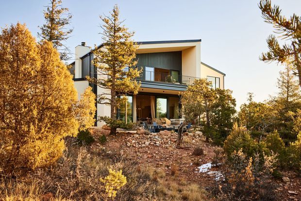 Exterior view of a contemporary two-storey home set within a dry, rocky, landscaped site. The house has a simple pale rendered facade, dark low-pitched roof, deep recessed upper balcony, dark metal balcony railings, large glazed openings, and a sheltered outdoor living area. Native and established trees surround the building, with sparse vegetation, exposed rocks, and a small patch of snow in the foreground.