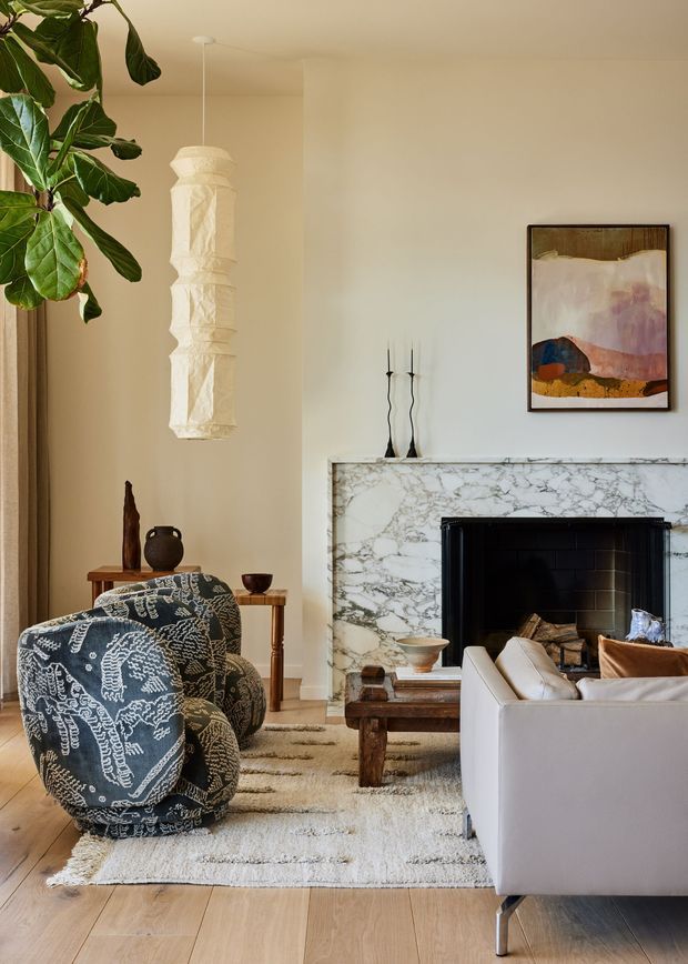 A warm, refined living room centered on a marble-clad fireplace with a black firebox. The room features a cream sofa, patterned accent armchair, timber coffee table, pale timber flooring, a textured neutral rug, a sculptural pendant light, indoor greenery, and abstract wall art. The overall composition is calm and contemporary with natural materials and soft beige tones.