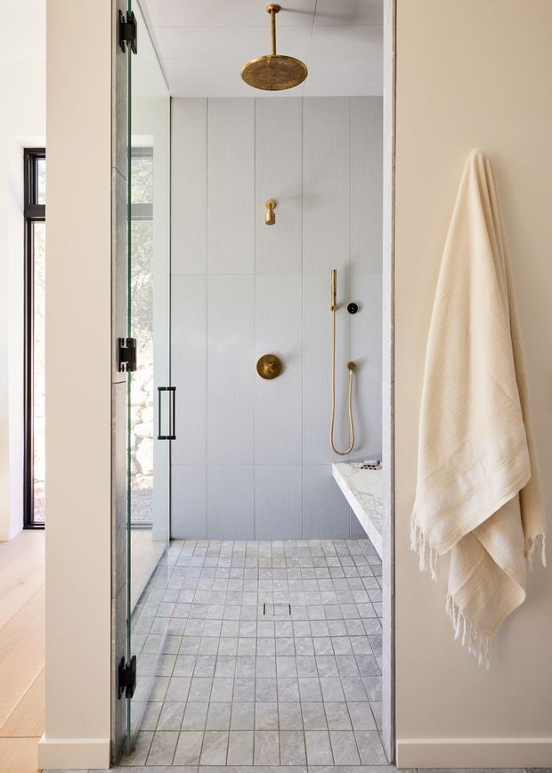 A narrow contemporary walk-in shower bathroom with a frameless glass enclosure, pale vertically panelled or large-format tiled walls, small square grey floor tiles, and brushed brass shower fixtures. A white stone bench projects from the right wall, while a soft cream towel hangs on the adjacent warm neutral wall. A slim window and light timber flooring are visible outside the shower area.
