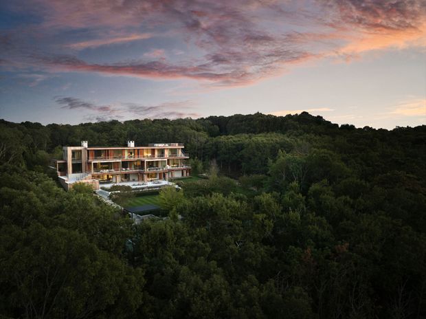 Aerial exterior view of a contemporary multi-storey residential building nestled within dense forested landscape. The long, rectilinear structure has extensive glazing, warm timber-toned elements, balconies, and a flat roof, with a visible pool or dark water feature and landscaped grounds below. The scene is photographed at dusk beneath a dramatic pink and blue clouded sky.