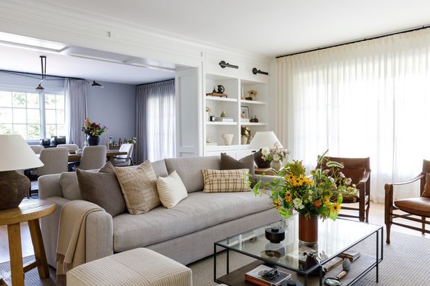 Bright, refined living room with a large upholstered sofa, layered neutral cushions, glass coffee table, armchairs, floral arrangement, built-in open shelving, and expansive sheer-covered windows. White wall panelling and trim create a classic architectural backdrop, while the overall scheme is soft, elegant, and inviting.