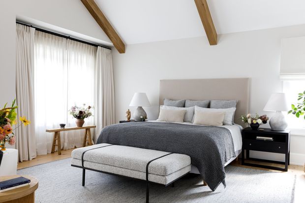 Bright, refined bedroom with a large upholstered bed, layered neutral bedding, a light grey upholstered bench, black bedside tables, sheer floor-length curtains, and exposed timber ceiling beams. White walls, pale flooring, and soft natural light create an airy contemporary interior.