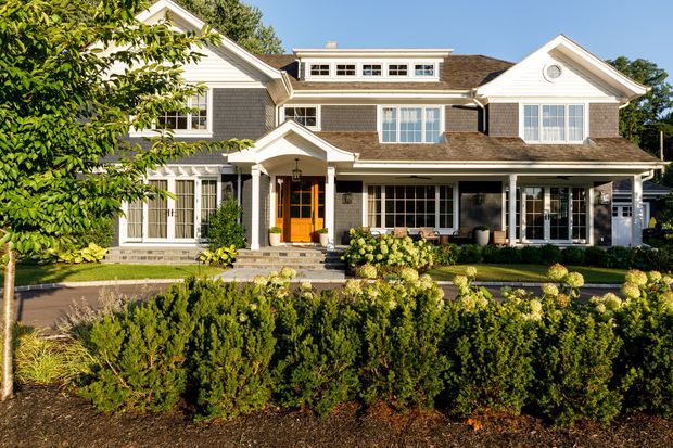 A large, symmetrical two-storey suburban residence with a traditional gabled roof, grey shingle-style exterior cladding, white architectural trim, multiple dormer-like upper windows, and a covered front porch. The home is set behind manicured hedges, shrubs, and planted borders, with a paved driveway or walkway leading to the entrance. Bright daylight highlights the formal, established landscaping and traditional facade.