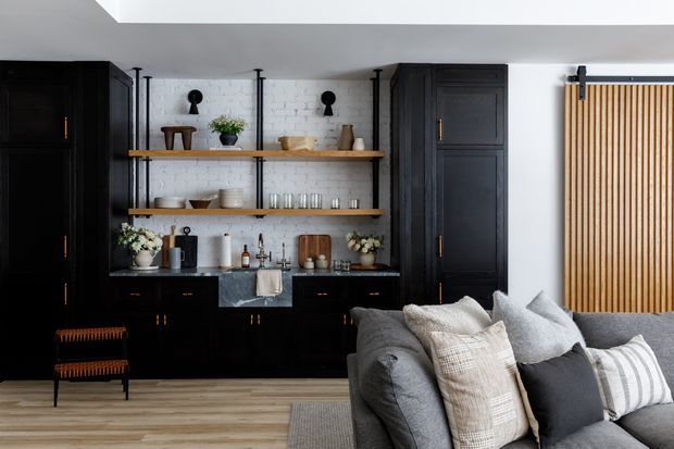A refined open-plan living area featuring a dark built-in kitchen or bar joinery wall with black cabinetry, brass hardware, open timber shelves, and a white painted brick backsplash. The space includes a grey sectional sofa with layered neutral cushions in the foreground, pale timber flooring, a black-and-tan accent chair, and a vertical timber slat screen at right. A large ceiling opening or skylight brings in natural light.