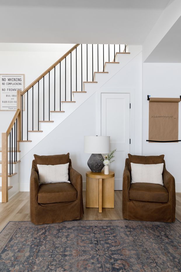 A bright, refined living room or sitting area with two brown slipcovered armchairs, white cushions, a round timber side table, and a sculptural lamp. A white staircase with slim black vertical balusters and a timber handrail rises behind the seating, with a white panel door beneath it. A patterned vintage-style rug spans the foreground, and framed wall art and a hanging scroll add understated decor.
