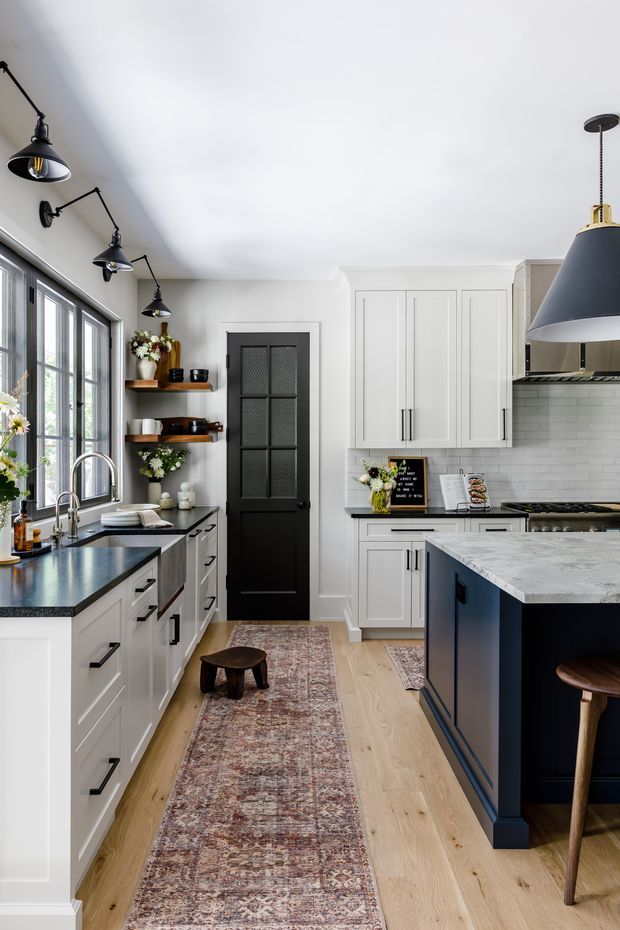 A bright, narrow contemporary kitchen with white Shaker-style cabinetry, dark navy island cabinetry, pale wood flooring, and dark stone countertops. The room features large black-framed windows, a black paneled door, open timber shelves with decor, black industrial-style wall lights, and a large dark pendant over the island. A patterned runner leads through the space, with a small stool and bar seating at the island.