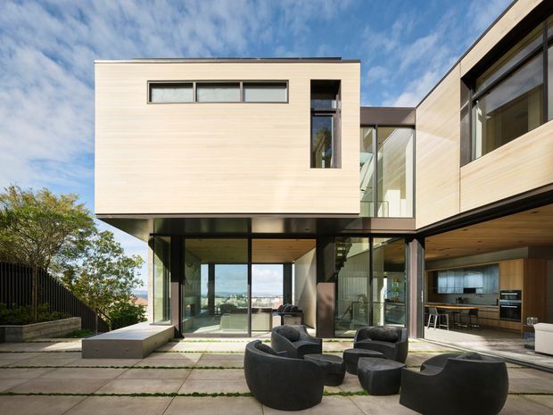 A contemporary multi-level residence viewed from an outdoor paved courtyard or terrace. The house features pale horizontal timber cladding, extensive floor-to-ceiling glazing, strong cantilevered forms, dark structural framing, and warm timber soffits. A partially visible open-plan kitchen is on the right, while the central glazing opens toward a distant urban or coastal outlook. Sculptural dark outdoor lounge chairs occupy the foreground.