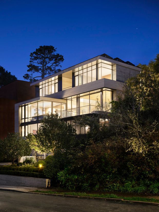 Twilight exterior view of a substantial multi-storey contemporary residence set among mature landscaped gardens. The building features extensive floor-to-ceiling glazing, projecting flat-roofed volumes, strong horizontal and vertical lines, illuminated interiors, and a mix of dark and light exterior surfaces. A street and retaining wall are visible in the foreground, with dense vegetation partially screening the lower levels.