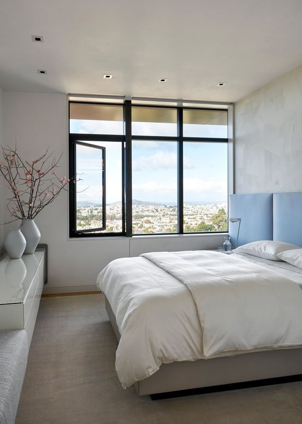 A bright, contemporary bedroom with a large black-framed window wall overlooking a city and hills. The room features a neatly made white bed with a pale blue upholstered headboard, neutral carpet, a white built-in or low-profile dresser, minimalist bedside table, recessed ceiling lights, and a sculptural vase with bare branches. One window panel is open, bringing in a sense of natural ventilation and expansive views.