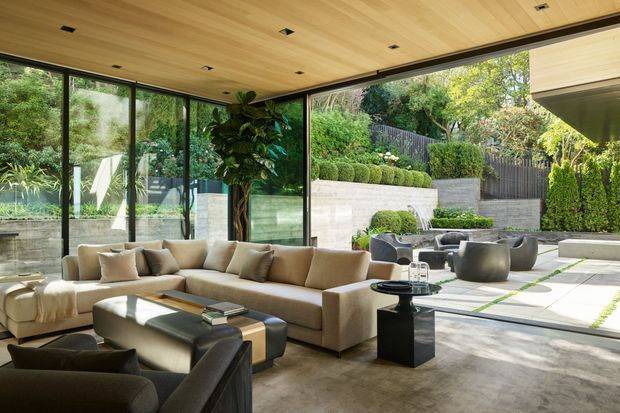 A spacious contemporary living room with floor-to-ceiling glazing opening onto a landscaped courtyard. The room features a large neutral sectional sofa, dark lounge chairs, a low coffee table, polished concrete flooring, and a warm timber-lined ceiling with recessed downlights. Outdoor seating and layered planting are visible beyond the glass.