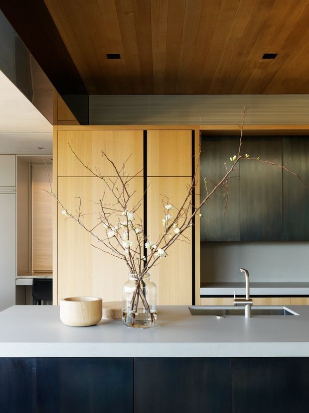 A refined contemporary kitchen viewed from the island. The space features a pale stone island benchtop with an undermount sink and slim chrome tap, dark blue-black lower cabinetry, tall light timber cabinetry with fine black reveals, and dark upper cabinetry. A warm timber-lined ceiling and horizontal textured wall panel add architectural warmth. Minimal branches in glass vessels provide a natural decorative accent.