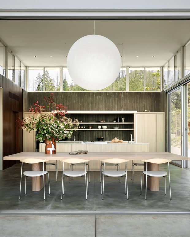 A refined open-plan kitchen and dining space with a long pale timber dining table, sculptural light-coloured chairs, and a large spherical pendant light. The kitchen features full-height dark timber or wood-look wall panelling, open shelving, pale built-in cabinetry, and a light benchtop. Floor-to-ceiling glazing on multiple sides brings in abundant natural light and views of trees. The space has a clean, minimalist architectural character with strong indoor-outdoor connection and carefully composed floral styling.