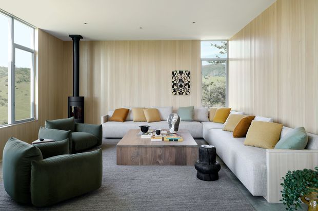 A bright contemporary living room with extensive floor-to-ceiling glazing overlooking a green landscape. Pale vertical timber wall panelling and a matching ceiling create a warm, natural backdrop for a large L-shaped light sofa, deep green lounge chairs, mustard and cream cushions, and a low timber coffee table. A black freestanding wood-burning stove and minimalist artwork provide contrasting focal points, while a large textured grey rug anchors the seating area.
