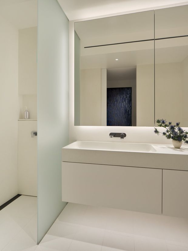Minimalist contemporary bathroom with a floating pale vanity, integrated white basin, large frameless mirrored cabinet, and concealed wall-mounted tapware. Light large-format floor and wall surfaces create a clean, bright interior, while a dark textured feature wall and small floral arrangement provide contrast.