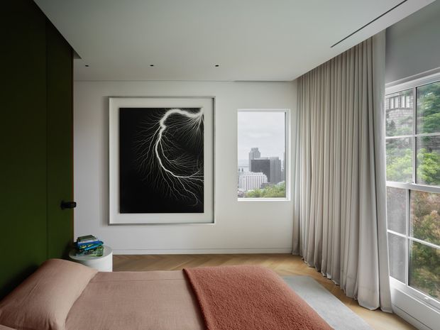 A refined contemporary bedroom with a low-profile bed in muted blush and terracotta textiles, pale timber flooring, white walls, and full-height sheer curtains. A dramatic black-and-white branching artwork forms the main feature, while large windows provide city views and abundant natural light. A dark green door or wall panel adds contrast.