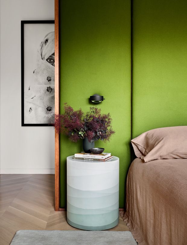 A close-up of a contemporary bedroom featuring a vivid lime-green vertically paneled feature wall, a partially visible bed dressed in taupe bedding, and a cylindrical layered pale green and white bedside table. A black wall sconce, framed monochrome artwork, flowers, books, timber trim, herringbone timber flooring, and a neutral rug complete the composed interior.
