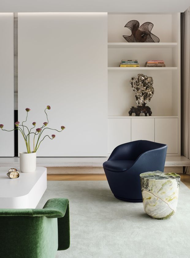 Refined contemporary living room with a sculptural navy lounge chair, green upholstered seating, a rounded white coffee table, and a veined stone side table. Built-in white shelving displays decorative sculpture and books, while a large white panelled feature wall and soft neutral surfaces create a polished, gallery-like setting.