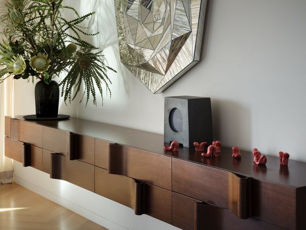 A contemporary interior vignette featuring a long floating dark timber sideboard with sculptural curved-front drawers and doors. A black vase with dried foliage, a compact speaker, and small red decorative objects sit on top. The white wall is accented by a large geometric mirrored artwork, while warm timber flooring and soft natural light create a refined, warm atmosphere.