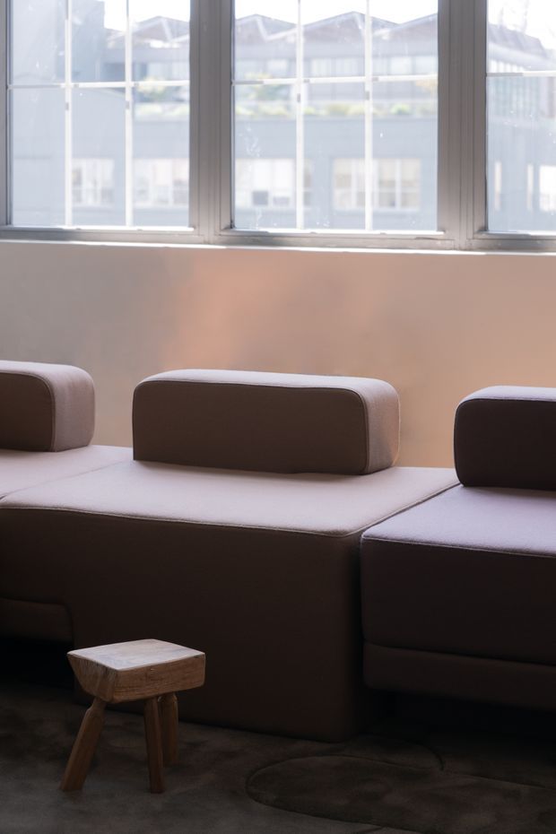 A softly lit contemporary living area featuring a large modular upholstered sofa in muted mauve-grey tones, positioned beneath tall industrial-style multi-pane windows. A small sculptural wooden side stool sits on a dark carpet, while warm sunlight creates a calm, intimate atmosphere.