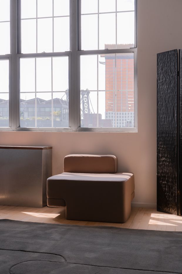 A softly lit contemporary interior corner featuring a large industrial-style multi-pane window overlooking an urban skyline. A sculptural brown upholstered lounge chair sits beside a dark built-in cabinet, with a tall black textured panel or screen along the right wall. Pale timber flooring and a large grey rug complete the calm, warm-toned setting.