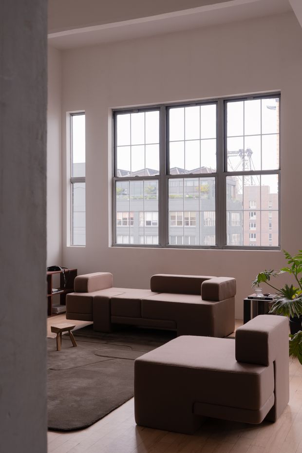 A contemporary living room viewed from an adjacent opening, featuring a large dark-framed multi-pane window, pale walls, timber flooring, a muted dark rug, and modular brown upholstered seating with a matching foreground armchair. A small side table, low shelving, and indoor plants complete the understated urban interior.