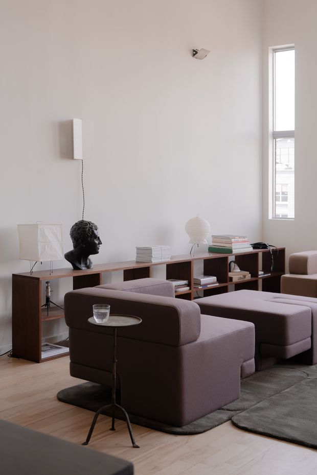 A refined living room with tall white walls, a narrow vertical window, pale timber flooring, and a long dark timber open-shelving console. Two upholstered mauve-grey lounge chairs and matching ottomans sit on layered grey rugs, accompanied by a slender metal side table. Sculptural table lamps, a wall-mounted light, books, and a dark bust add understated decorative detail. The room has a clean, calm, contemporary feel with generous vertical proportions.